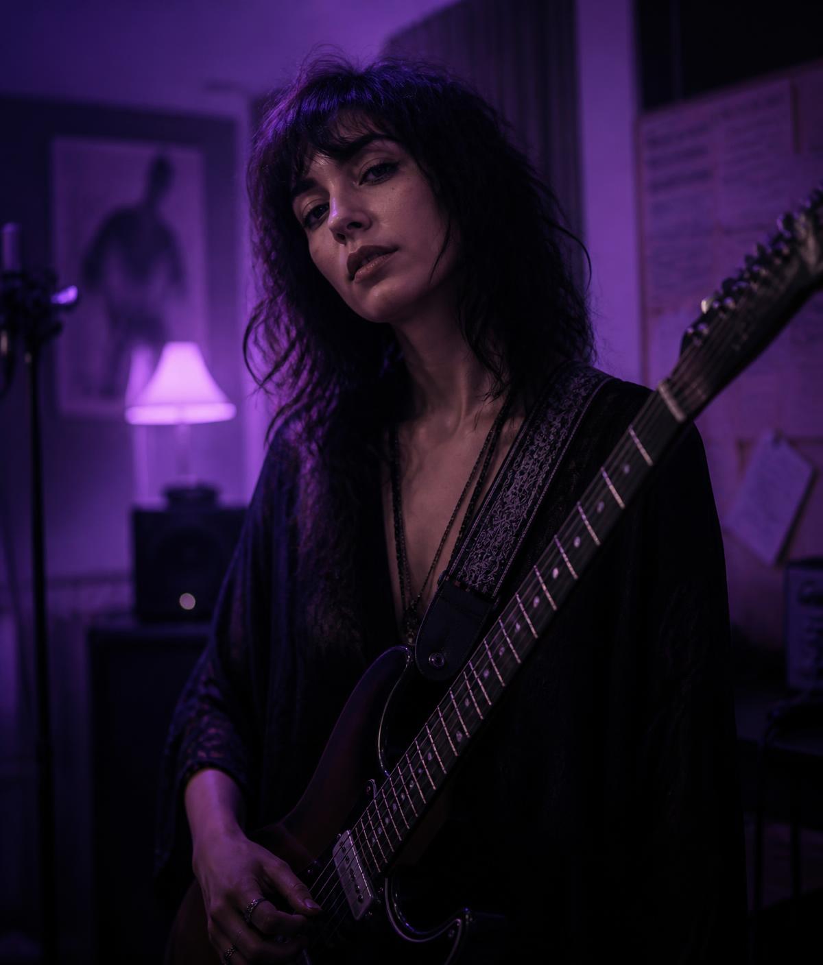 An emerging solo artist holding a guitar in a dimly lit rehearsal room with soft lilac lighting